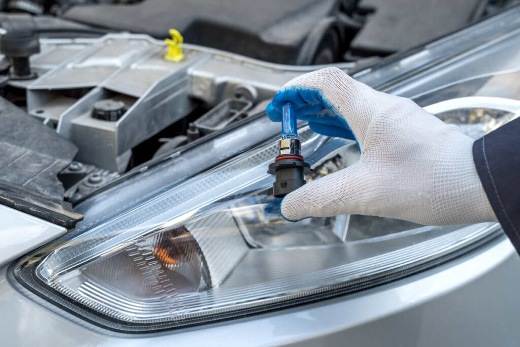mano con guanto che tiene una lampadina per faro davanti ad un cofano di un auto aperto